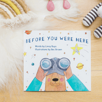 Children's book titled 'Before You Were Here' on a wooden surface with toys around