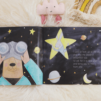 Illustrated book open to a page with a bear looking through binoculars and a star-shaped rabbit, on a soft surface.
