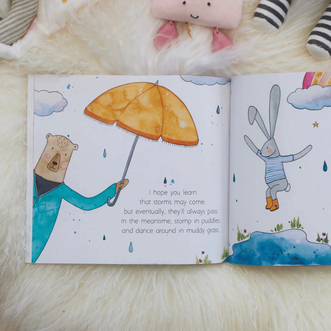 Open children's book with illustrations of a bear holding an umbrella and a rabbit in the rain, on a soft surface.