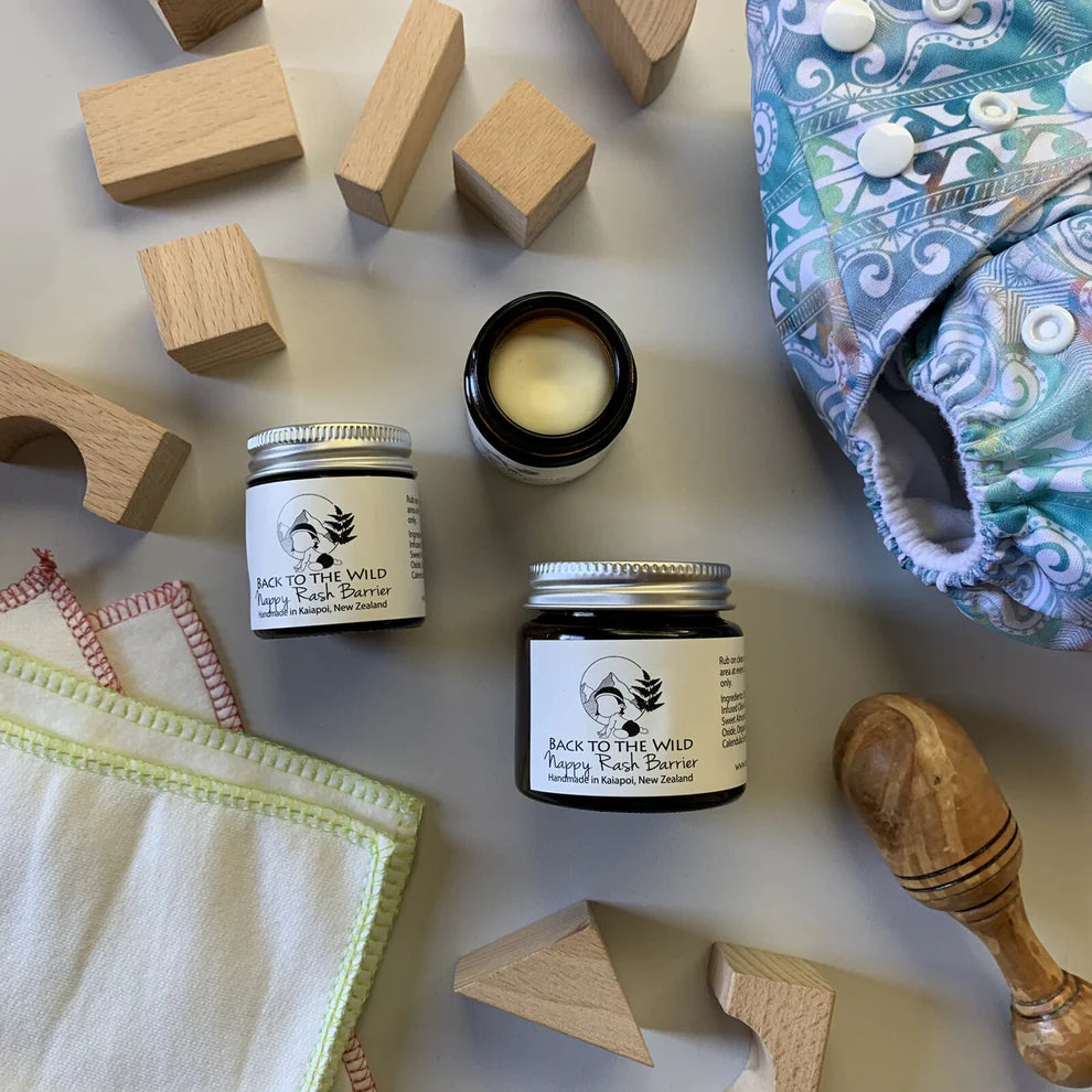 The image shows a small open jar of 'Nappy Barrier Balm' with its contents partially visible, placed next to a wooden block and a cloth with a pattern. In the background, there's a diaper and a wooden spoon lying on the surface.