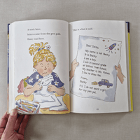 Open book showing a page with an illustration of a girl and text, held by a hand on a textured surface.