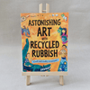 Book titled 'Astonishing Art with Recycled Rubbish' on a wooden easel against a grey background