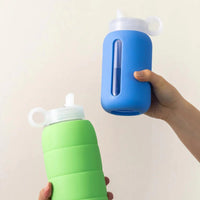 Two glass water bottles with silicone covers, one green and one blue, held by a hand against a plain background.