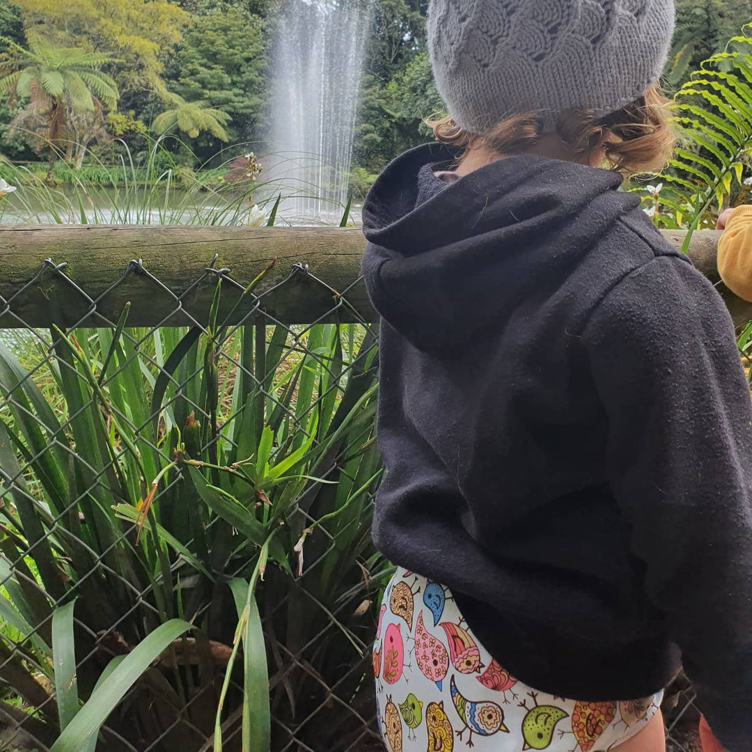 Child wearing reusable nappy, a grey knit hat and dark hoodie looking at a fountain in a park.