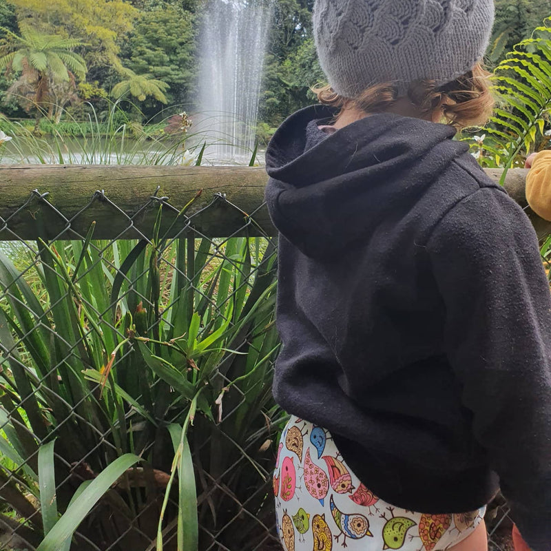 Child wearing reusable nappy, a grey knit hat and dark hoodie looking at a fountain in a park.