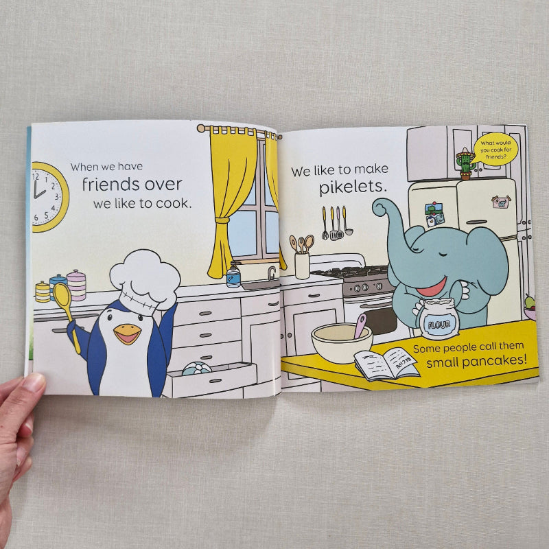 Open children's book with cartoon characters in a kitchen setting on a gray surface.