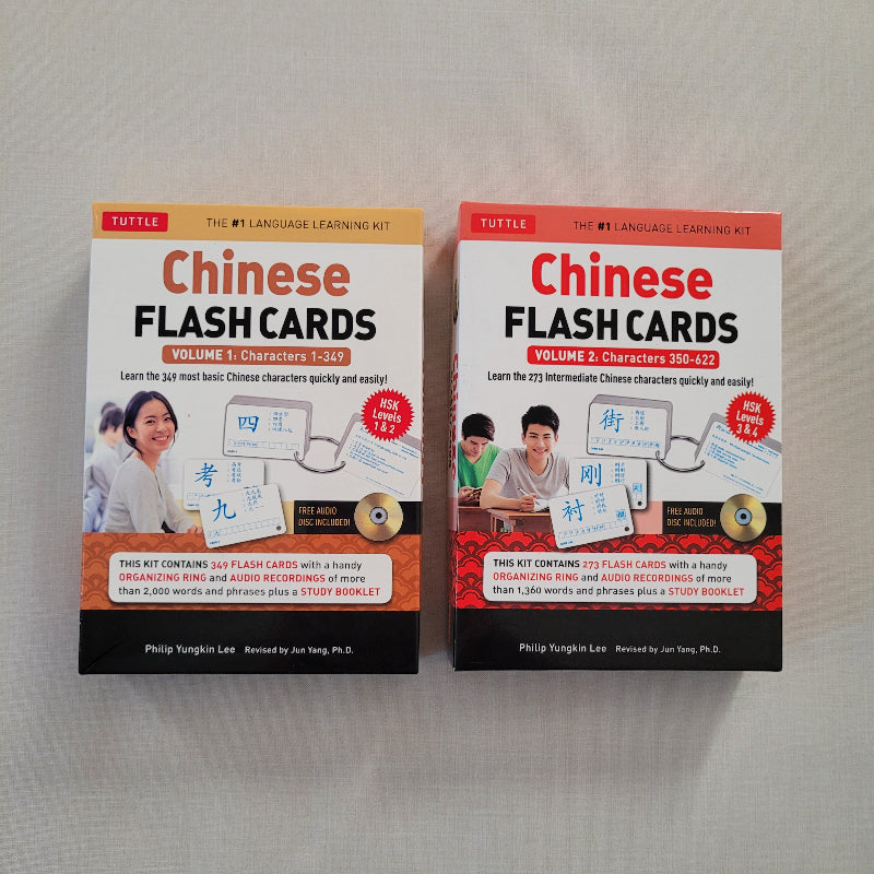 Two boxed Chinese Flash Cards learning kits by Tuttle, shown side by side on a light fabric background. The left box is Volume 1 with 360 flash cards, and the right box is Volume 2 with 720 characters, both with orange, white, and red packaging and photos of learners on the front.