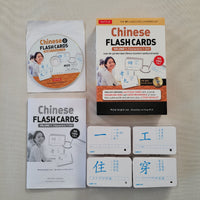 Chinese Flash Cards Volume 1: Characters 1-148 language learning kit by Tuttle, including the main book, a CD, a booklet, and four sample flashcards with Chinese characters, arranged on a light beige fabric background.