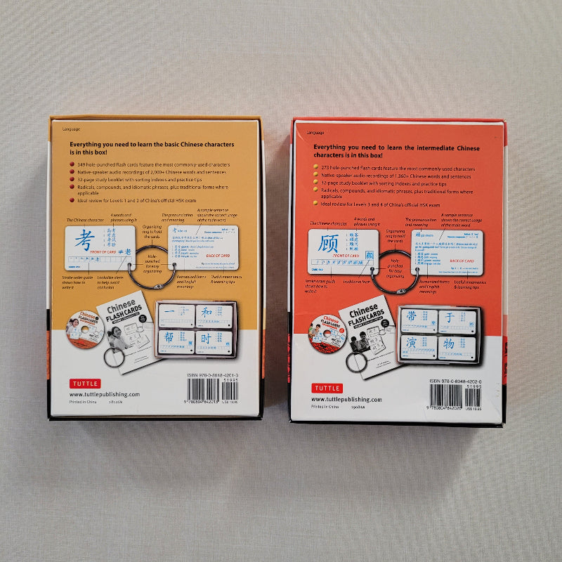 Two boxed Chinese language learning products shown side by side on a light gray fabric background. The left box is tan and the right box is coral-red, both showing the back covers with text, diagrams, and product imagery.