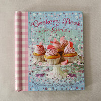 Cookery book titled 'Cookery for Girls' with a colourful cover featuring cupcakes and floral patterns on a textured surface.