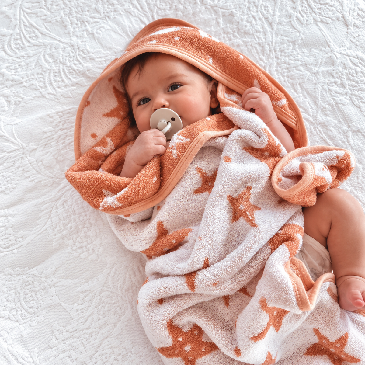 Baby wrapped in cotton hooded bath towel with coral orange and white coloured starfish on white patterned background
