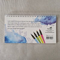 Spiral-bound notebook with watercolor design and text on a gray fabric background