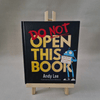 Book titled 'Do Not Open This Book' by Andy Lee on a stand with a grey background