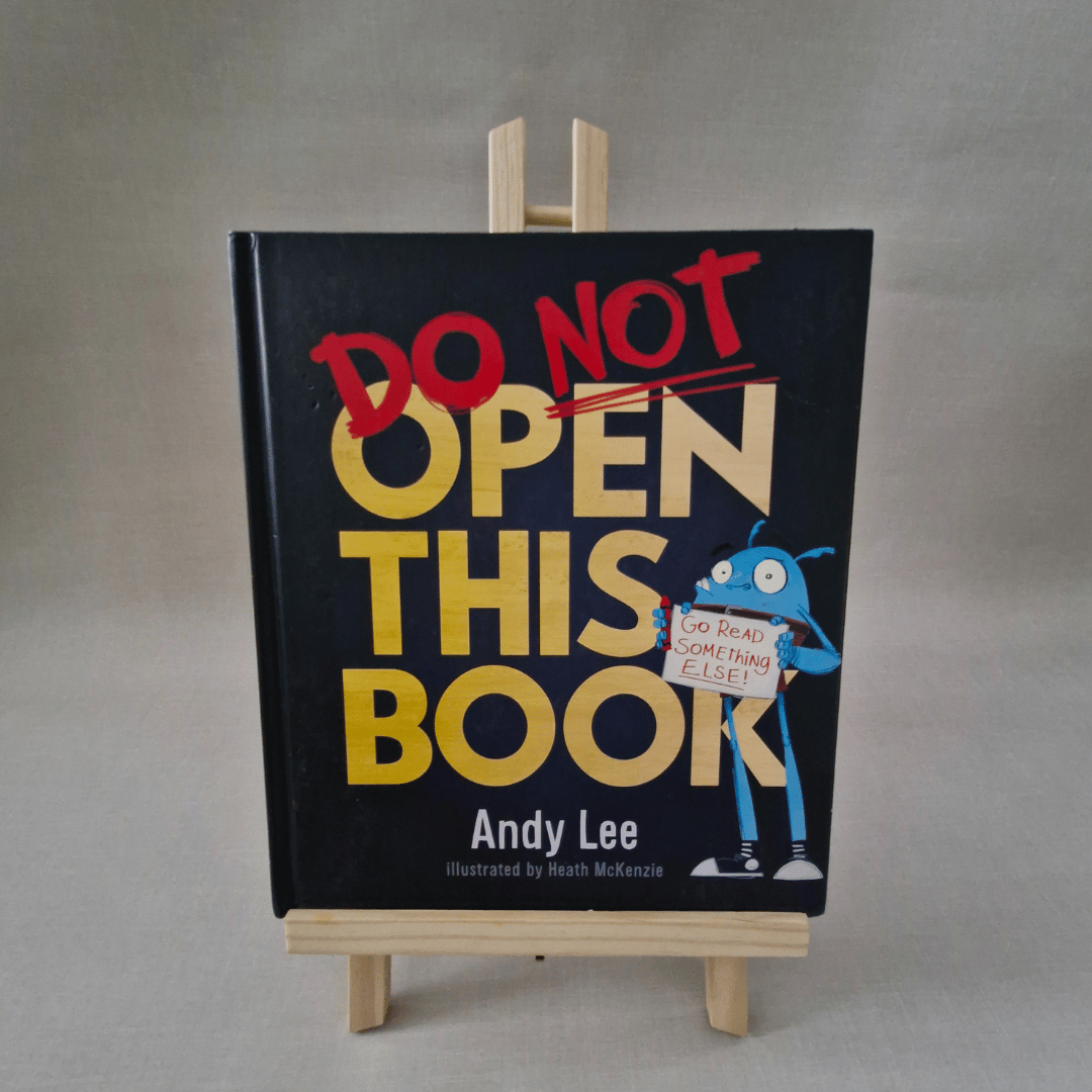 Book titled 'Do Not Open This Book' by Andy Lee on a stand with a grey background