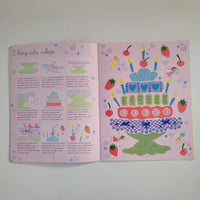 An open pink booklet or collage page showing colorful illustrated fairy cake designs and small printed text panels, photographed against a plain light background.