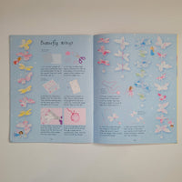 An open book on a light beige surface. The pages are pale blue and decorated with many pastel butterfly illustrations in pink, yellow, white, and blue, plus small step-by-step sewing or craft instructions and a title reading 'butterfly strings.