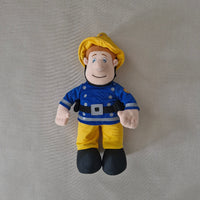Plush toy figure of a character in a blue shirt and yellow pants on a grey background