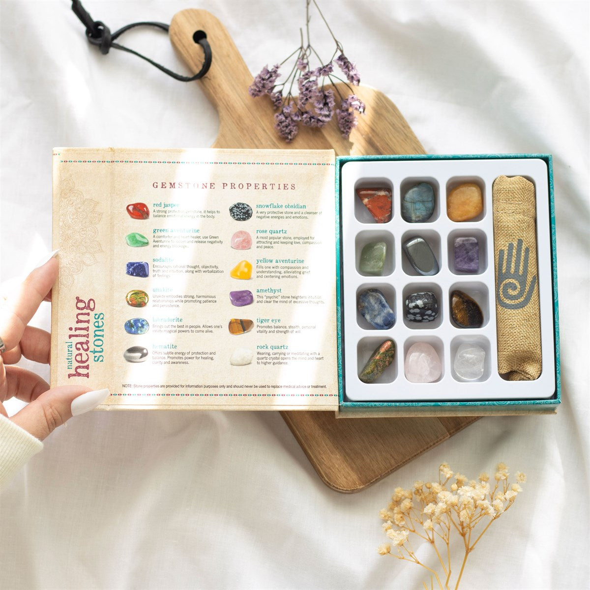 Open box showing 12 natural gemstones in separate slots with jute bag and description of each stone, resting on a chopping board with dried flowers and white fabric background