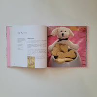 An open recipe book laid flat on a light neutral surface, showing a printed page with text and a photo of a bone shaped cookie in a basket for dogs.
