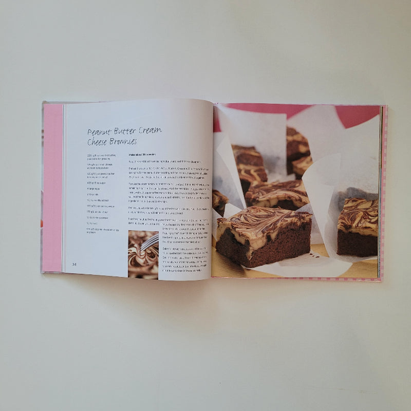 An open cookbook on a light beige surface, showing a page with a photo of layered dessert bars and printed recipe text. The book has pink side borders and a white cover edge visible.