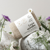 Two eco scrubs with a label on a stone surface surrounded by flowers and plants.