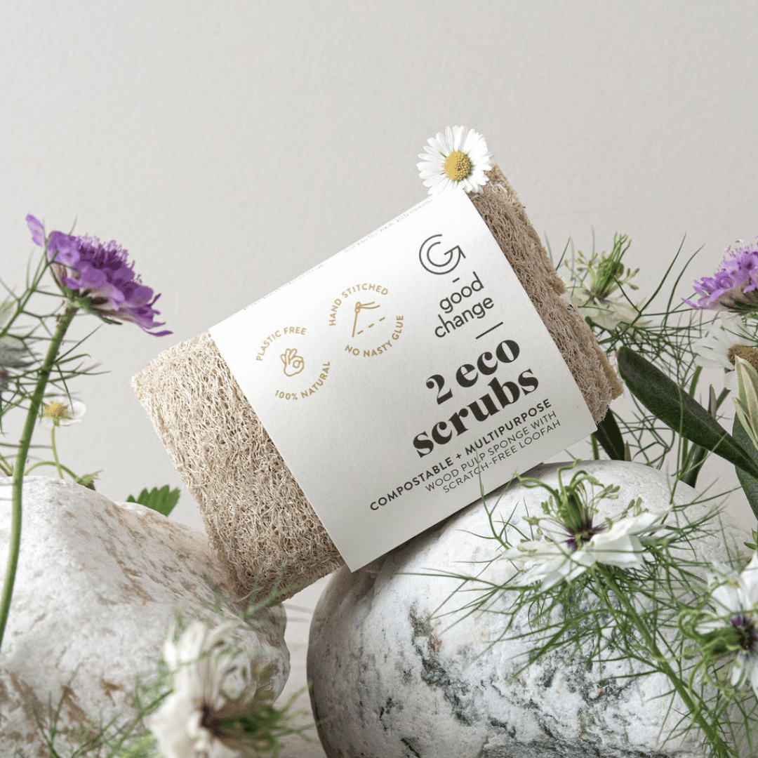 Two eco scrubs with a label on a stone surface surrounded by flowers and plants.