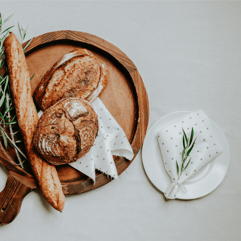 Loaves of bread on a wooden board with a reusable bamboo towel as a napkin and greenery on a light gray background
