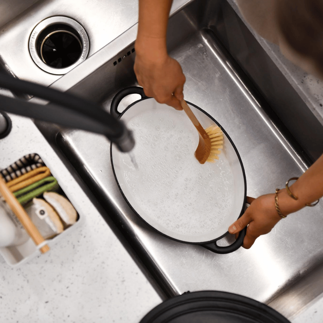 Person washing a black pot with a wooden scrubber in a kitchen sink.