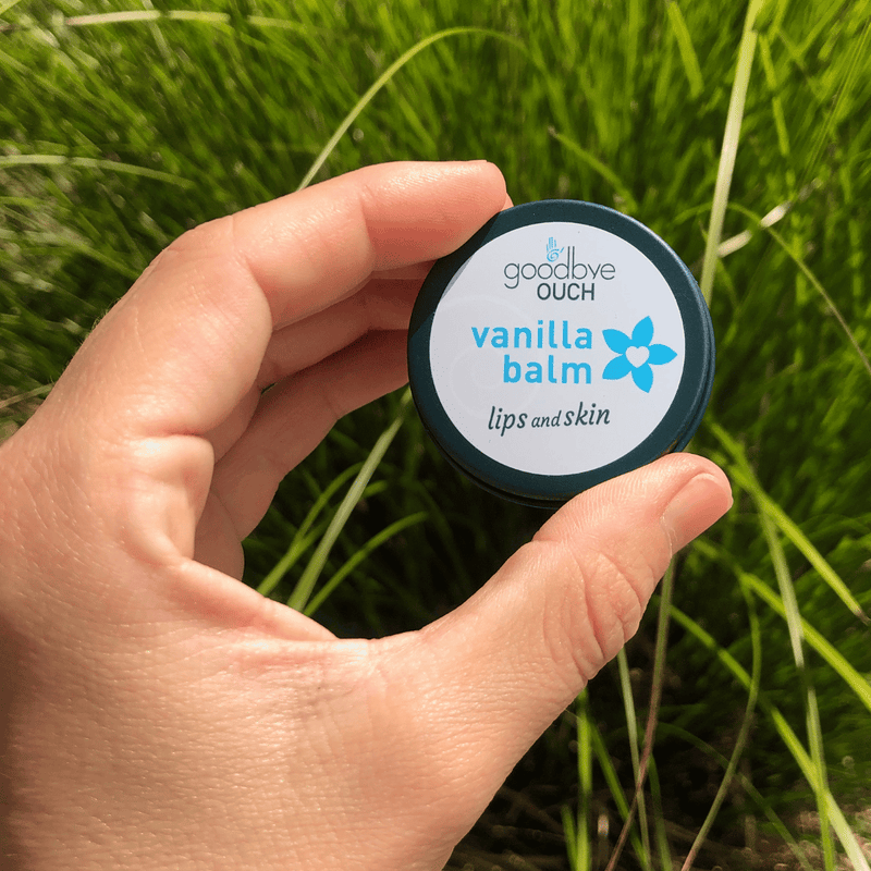 Hand holding a container of 'Goodbye Ouch Vanilla Balm' against a grassy background