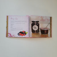 An open spiral-bound  cookbook laid flat on a light neutral background. The left page shows a glass jar filled with dark jam alongside a small bowl, and the right page contains a recipe with a berry illustration and decorative pink swirls.