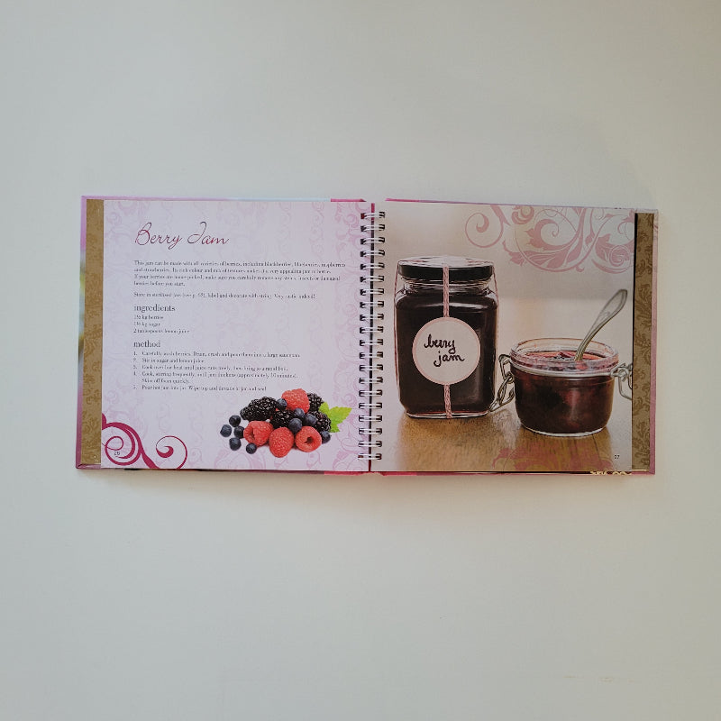 An open spiral-bound  cookbook laid flat on a light neutral background. The left page shows a glass jar filled with dark jam alongside a small bowl, and the right page contains a recipe with a berry illustration and decorative pink swirls.