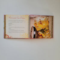 An open spiral-bound cookbook showing a colourful food photograph of marinated feta cheese on one page and printed recipe text on the other.