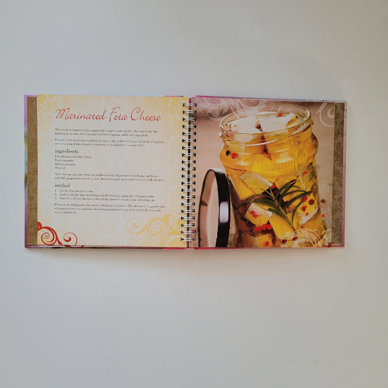 An open spiral-bound cookbook showing a colourful food photograph of marinated feta cheese on one page and printed recipe text on the other.
