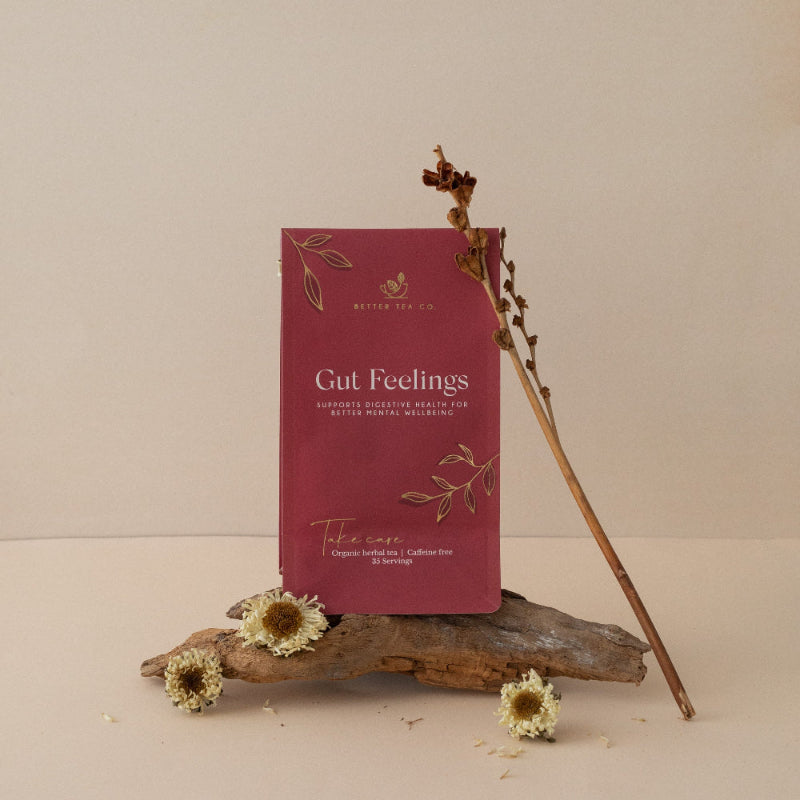 A package of 'Gut Feelings' herbal tea in a compostable pouch, accompanied by dried flower accents and a branch, placed on a wooden surface with a beige background.
