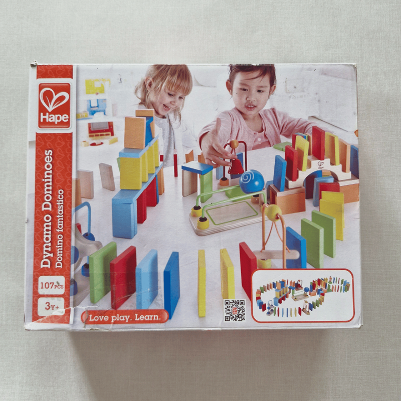 Hape Dynamo Dominoes toy packaging with two children playing with the product on a light grey background.
