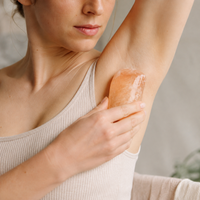 A woman using a pinkish-orange himalayan salt deodorant bar on her underarm, wearing a ribbed beige tank top.