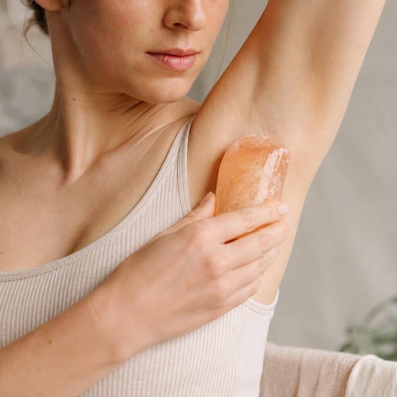 A woman using a pinkish-orange himalayan salt deodorant bar on her underarm, wearing a ribbed beige tank top.