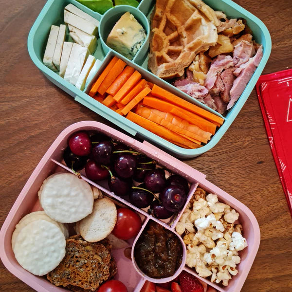 Two bento boxes filled with a variety of snacks including carrots, cheese, and fruits on a wooden surface.