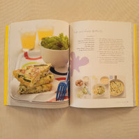An open cookbook spread on a beige surface, showing printed recipe pages on the left and a plated breakfast photo on the right with a fork, omelette, eggs, and greens.