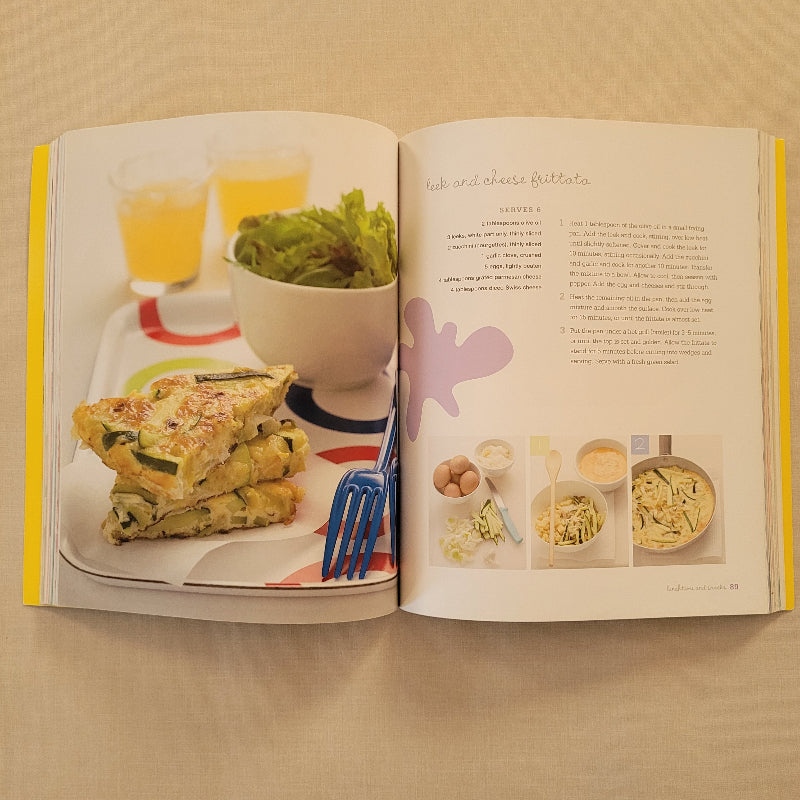 An open cookbook spread on a beige surface, showing printed recipe pages on the left and a plated breakfast photo on the right with a fork, omelette, eggs, and greens.