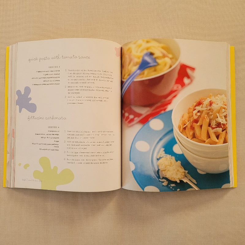 An open cookbook laid flat on a beige surface, showing two interior pages with a food photograph of pasta dishes on the left page and recipe text on the right page.