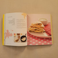 An open printed recipe book spread showing the preparation of quesadillas, with a photo of stacked pile on a plate on the right page and ingredients on the left page, set against a beige textured background.