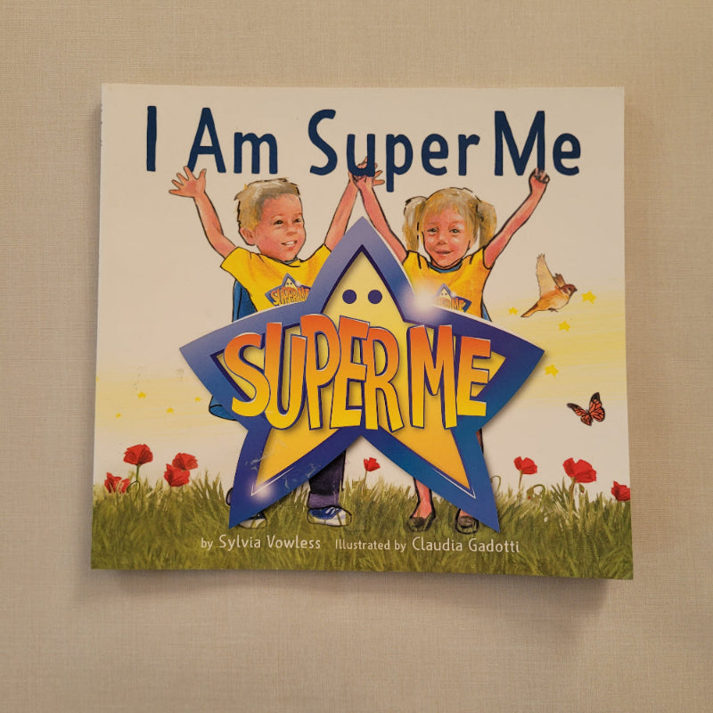 A children's book titled 'I Am Super Me' by Silvia Nowles, featuring a colorful cover illustration of two children in yellow shirts with a large blue and yellow star graphic, photographed on a beige fabric background.