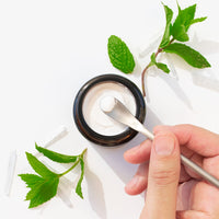 Hand using a scoop to take toothpaste from a jar with green mint leaves on a white background