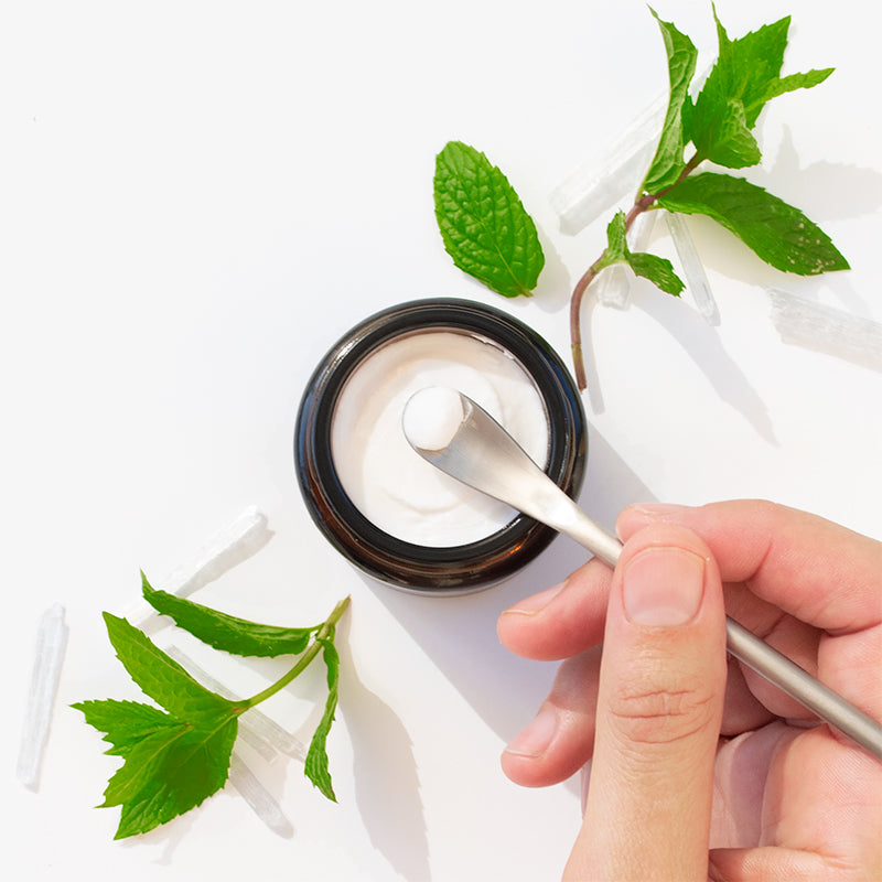 Hand using a scoop to take toothpaste from a jar with green mint leaves on a white background