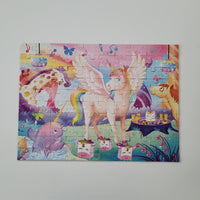 A completed children's jigsaw puzzle featuring a whimsical unicorn party scene, displayed flat against a light gray wall. The puzzle shows a pink winged unicorn, a purple unicorn, a pink pig, and colorful musical notes and flowers.