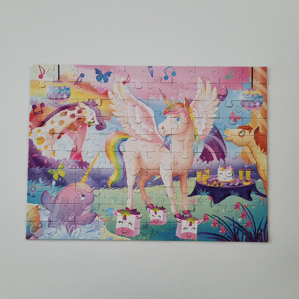 A completed children's jigsaw puzzle featuring a whimsical unicorn party scene, displayed flat against a light gray wall. The puzzle shows a pink winged unicorn, a purple unicorn, a pink pig, and colorful musical notes and flowers.