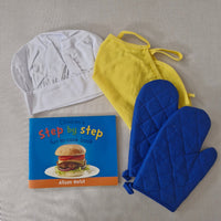 Yellow apron, blue oven mitts, white chef hat, and 'Step by Step' book on a gray surface