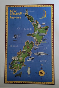 A completed jigsaw puzzle featuring a colourful illustrated map of New Zealand, with the title 'New Zealand Aotearoa' at the top, surrounded by a decorative border and mounted on a light wall.