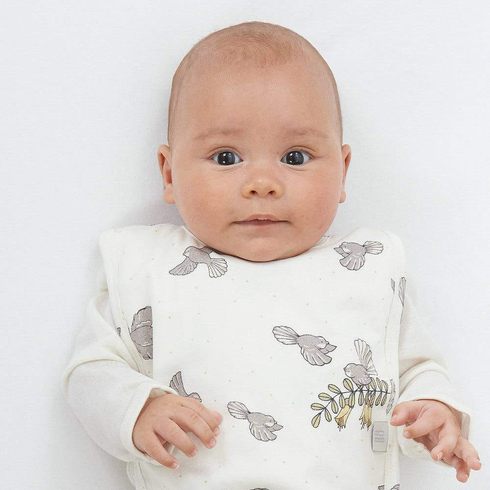 Baby wearing a white outfit with fantail piwakawaka patterns on a plain background